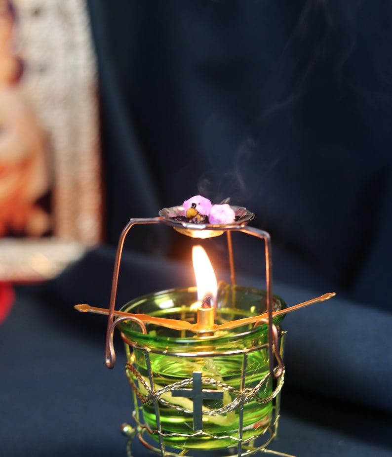 May include: A lit green glass candle holder with a cross design, surrounded by a gold wire cage. A small metal dish above the flame holds purple flowers. Smoke rises from the flame against a dark background.