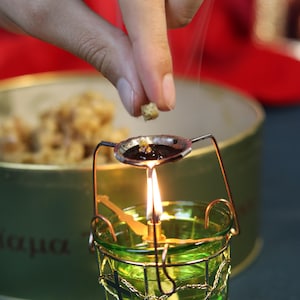 May include: A lit green glass candle holder with a metal frame and a small spoon-like dish above the flame. A hand is adding a small piece of material to the dish. A container of material is in the background.
