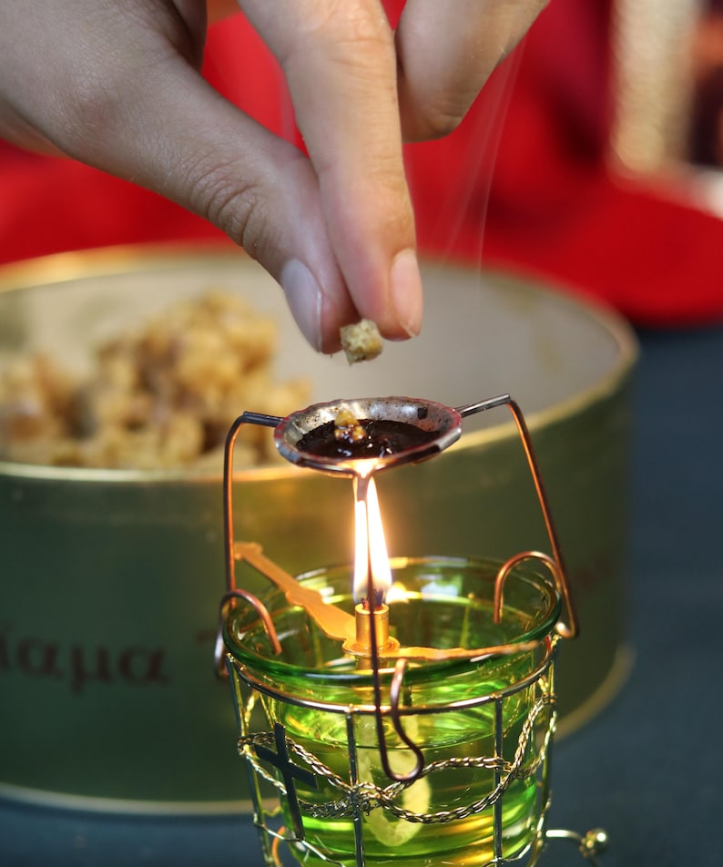 May include: A lit green glass candle holder with a metal frame and a small spoon-like dish above the flame. A hand is adding a small piece of material to the dish. A container of material is in the background.