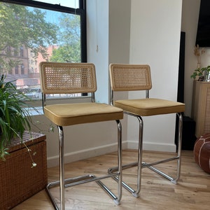 May include: Two chrome bar stools with woven cane backs and mustard yellow velvet seats. The stools are on a light wood floor.