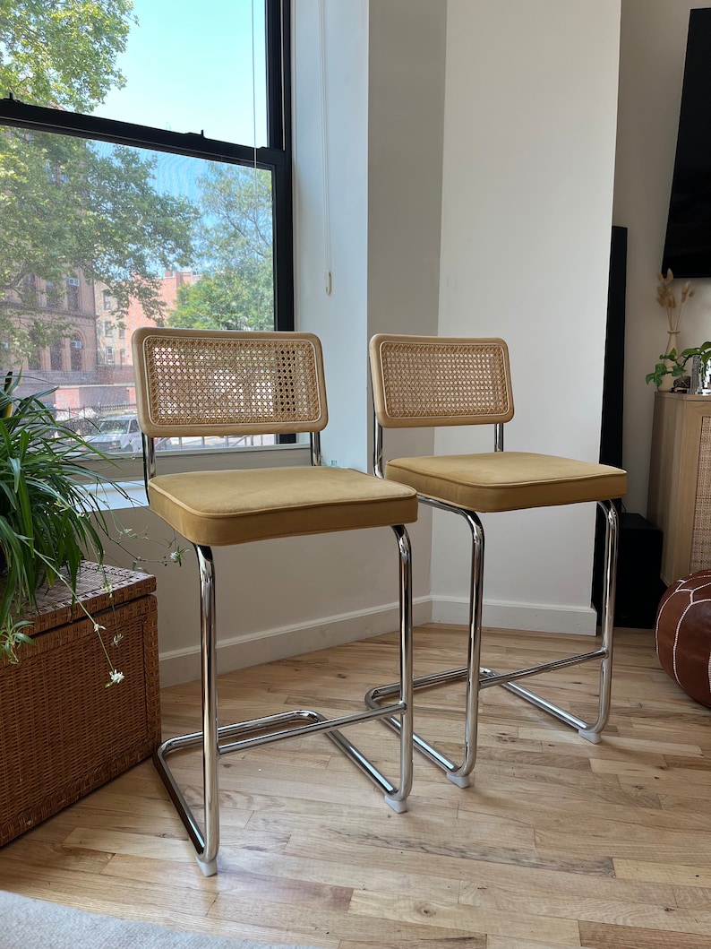 May include: Two chrome bar stools with woven cane backs and mustard yellow velvet seats. The stools are on a light wood floor.