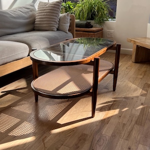 May include: Mid-century modern coffee table with a glass top and a lower shelf made of woven rattan. The table has a dark wood frame and is oval-shaped. The design is a blend of wood, glass, and natural materials.