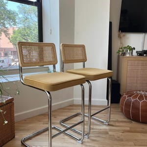 May include: Two chrome bar stools with woven cane backs and beige upholstered seats. The stools are on a wooden floor.