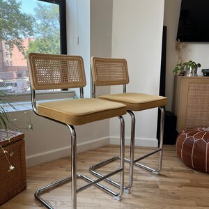 May include: Two chrome bar stools with tan velvet seats and woven cane backs. The stools are on a hardwood floor.