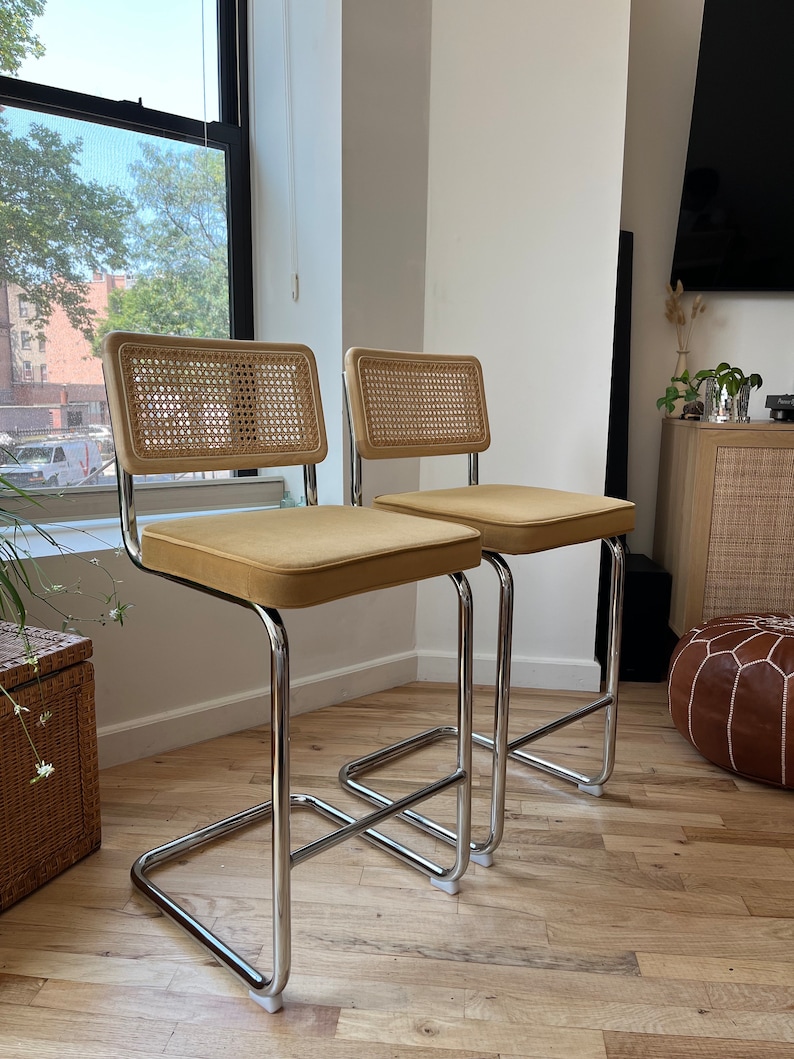 May include: Two chrome bar stools with tan velvet seats and woven cane backs. The stools are on a hardwood floor.