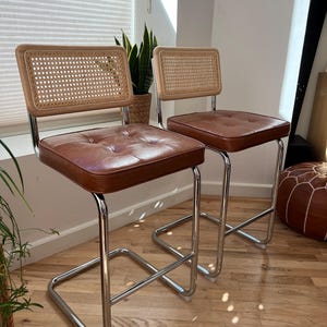 May include: Two brown leather bar stools with chrome frames and woven cane backs. The stools are on a wooden floor.