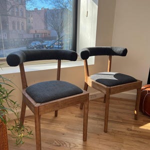May include: Two wooden chairs with dark gray fabric seats and backs. The chairs have a unique curved back design and are set on a wooden floor.