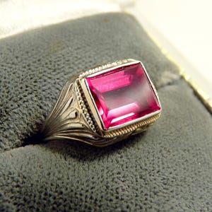 May include: A silver-toned ring featuring a large, rectangular, faceted deep pink gemstone set in a decorative metal frame with intricate side detailing and a beaded border surrounding the stone, showcasing a polished and reflective surface.
