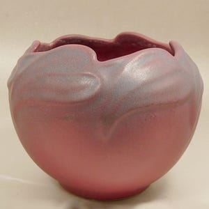 May include: A round, ceramic vase in a muted rose color with a ruffled rim. The vase has a smooth, glossy finish with subtle, swirling patterns in a lighter, silvery hue near the top. The overall shape is organic and flowing.