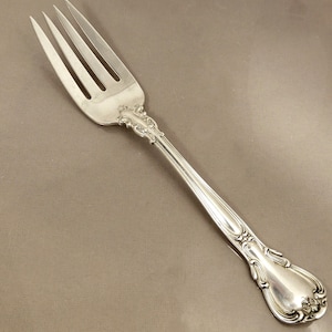May include: A silver fork with a decorative handle. The fork has four tines and a rounded handle with ornate details.
