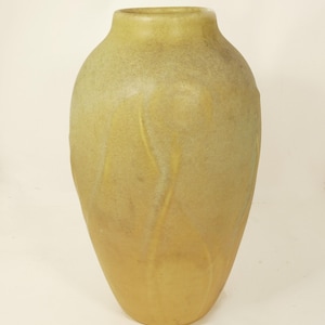 May include: A tall, bulbous ceramic vase with a matte finish in shades of muted yellow, ochre, and soft green. The surface features subtle, raised vertical wave-like patterns that wrap around the body, leading to a narrow, circular opening.