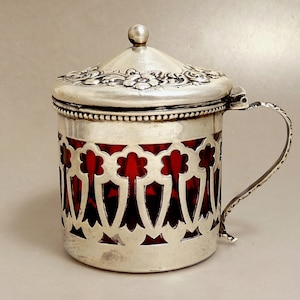 May include: A silver-colored cup with a handle and a lid. The cup's body features a decorative openwork design revealing a red interior. The lid is adorned with floral patterns and a small spherical knob. The handle is curved and detailed.