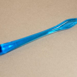 May include: A long, curved, blue glass rolling pin with a rounded handle and a flattened end. The glass is transparent, allowing light to pass through, and has a smooth, glossy finish. The rolling pin is designed for kitchen use.