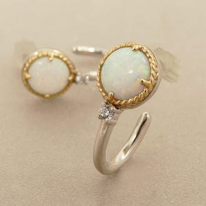May include: A pair of opal earrings. Each earring features a round, white opal stone set in a gold-toned, rope-textured bezel. A small, clear gemstone is set on the silver-toned hook. The earrings are displayed on a neutral background.