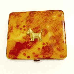 May include: A rectangular compact with a marbled pattern in shades of yellow and red. A gold-colored terrier dog is affixed to the top. The compact has a clasp closure and a shiny finish.