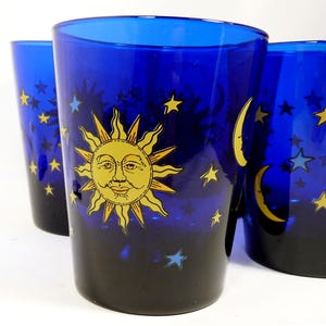May include: Set of three cobalt blue drinking glasses. Each glass features a celestial design with a yellow sun, crescent moons, and stars. The glasses have a gradient effect, transitioning from black to blue.
