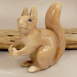 May include: A ceramic squirrel figurine in a light brown glaze. The squirrel is posed holding a nut, with a bushy tail curved upwards. The figurine has small blue eyes and detailed features.