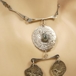May include: A silver-toned necklace with a textured, hammered design. The necklace features a large, circular pendant with a central stone and two smaller, irregularly shaped pendants. The metal has a slightly tarnished appearance.