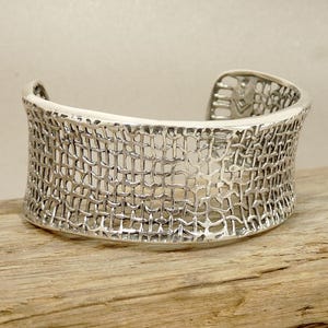 May include: A wide silver cuff bracelet featuring an intricate, openwork mesh pattern that resembles woven fabric. The metal has a polished, reflective finish, and the design is consistent across the entire curved surface of the cuff.