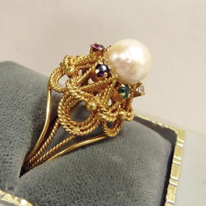 May include: A gold-toned ring featuring a large, round pearl as the focal point. The ring's design incorporates intricate, rope-like detailing and is adorned with small, colorful gemstones, including ruby, emerald, and amethyst accents. The ring is presented on a gray velvet surface.