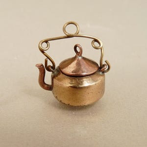 May include: A miniature copper-toned metal teapot charm featuring a rounded body, a small spout, and a conical lid with a central loop. The teapot has a decorative, arched wire handle with curled ends attached to the sides of the vessel.