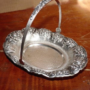 May include: A silver oval serving tray with a decorative handle. The tray has a hammered finish and intricate floral designs around the edge.