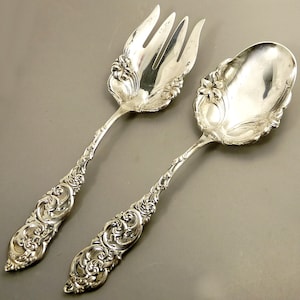 May include: A set of two silver serving utensils, a fork and a spoon, with ornate floral designs. The utensils are made of polished silver and have a vintage look.