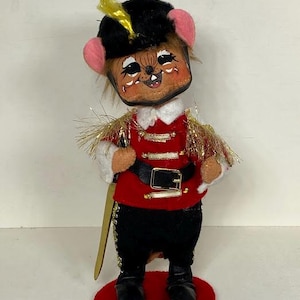 Puede incluir: Un juguete de peluche de ratón vestido como un soldado rojo y dorado. El ratón tiene un sombrero negro con una pluma amarilla, una chaqueta roja con ribetes dorados y pantalones negros. El ratón sostiene una espada dorada y está de pie sobre una base roja.