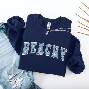 Pu&ograve; includere: Una felpa blu navy con la parola "Beachy" stampata in azzurro sul davanti. La felpa &egrave; piegata e appoggiata su una superficie bianca con un paio di jeans blu.