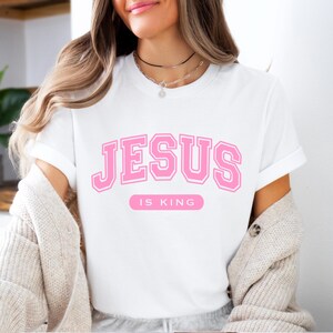 May include: White t-shirt with the text "JESUS IS KING" in pink block letters. The shirt has short sleeves and a crew neck. The text is in a collegiate style font.