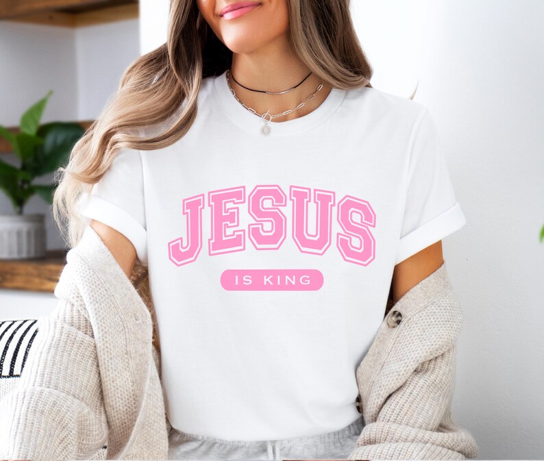 May include: White t-shirt with the text "JESUS IS KING" in pink block letters. The shirt has short sleeves and a crew neck. The text is in a collegiate style font.