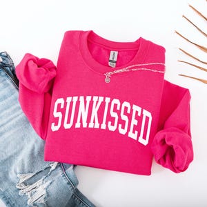 May include: A pink sweatshirt with the word "SUNKISSED" in white letters. The sweatshirt is folded and laid on a white surface with a pair of blue jeans.