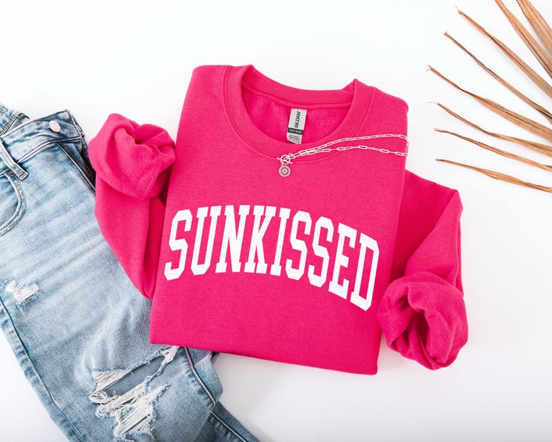 May include: A pink sweatshirt with the word "SUNKISSED" in white letters. The sweatshirt is folded and laid on a white surface with a pair of blue jeans.