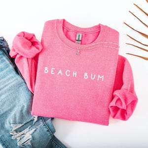May include: A pink sweatshirt with the text "Beach Bum" in white lettering. The sweatshirt is laid out on a white surface with a pair of blue jeans and a palm leaf.