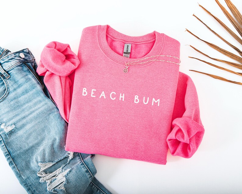 May include: A pink sweatshirt with the text "Beach Bum" in white lettering. The sweatshirt is laid out on a white surface with a pair of blue jeans and a palm leaf.
