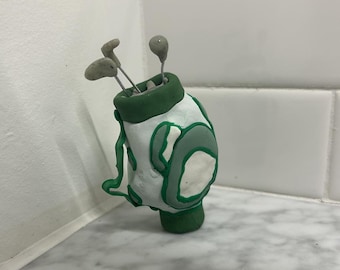 May include: A handmade golf bag sculpture in white, green, and gray. The bag has a green rim and base, with green accents and three golf clubs. The sculpture is made of clay and sits on a white and gray surface.
