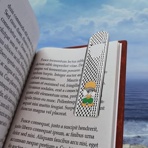 Pode incluir: Um marcador de livros decorativo com um padrão xadrez em preto e branco e um personagem de desenho animado. O marcador de livros está colocado num livro aberto. O fundo mostra um céu azul e o mar.