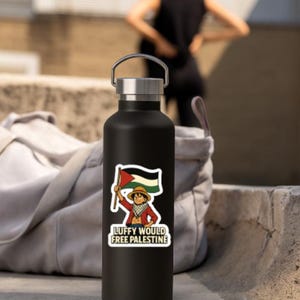 May include: Black water bottle with a silver lid and handle. A sticker on the bottle features a cartoon character holding a flag with the colors of the Palestinian flag, with the text "LUFFY WOULD FREE PALESTINE".