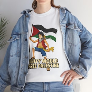 Può includere: Una t-shirt bianca con una grafica del personaggio anime Monkey D. Luffy che tiene in mano una bandiera palestinese. Sotto il personaggio, il testo recita LUFFY WOULD FREE PALESTINE in lettere scure. La maglietta è indossata sotto una giacca di jeans.