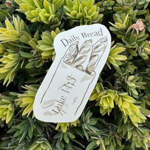 Könnte beinhalten: Ein Aufkleber mit einer Zeichnung von drei Baguettes in einer Papiertüte. Die Worte "Daily Bread" und "Luke 1:3" sind auf dem Aufkleber aufgedruckt. Der Aufkleber befindet sich vor einem Hintergrund aus grünem und gelbem Laub.