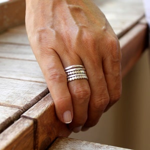 May include: A hand wearing a stack of silver rings. The rings have various designs, including hammered textures and beaded patterns. The rings are worn on the ring finger and middle finger.