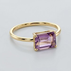 A close-up shot of a yellow gold ring featuring an emerald-cut purple and yellow ametrine stone against a white surface.