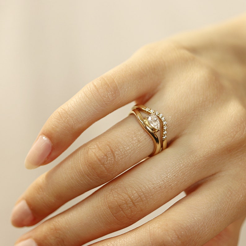 Pode incluir: Um anel de ouro com um diamante em forma de p&ecirc;ra e uma banda de diamantes menores.