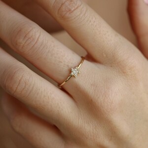 A woman's hand with light pink nail polish wears a thin gold ring with a small diamond-studded star on her ring finger, viewed from a slightly different angle.
