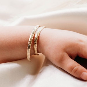 May include: Two gold bangle bracelets with an intricate floral design. The bracelets are worn on a child's wrist.
