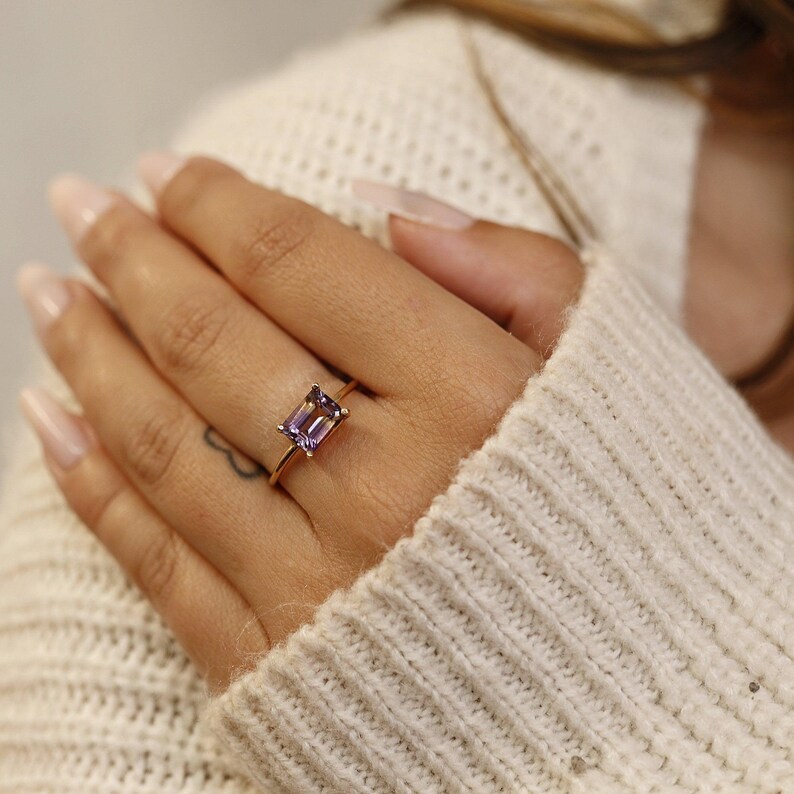 A hand wearing a gold emerald-cut ametrine ring, shown against a white knit sweater background.