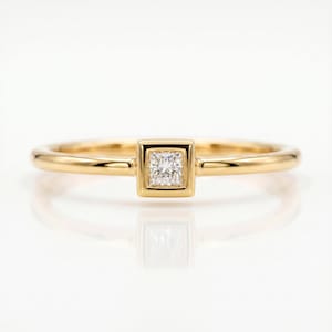 A gold ring with a bezel-set square princess cut diamond centered on a white reflective surface.
