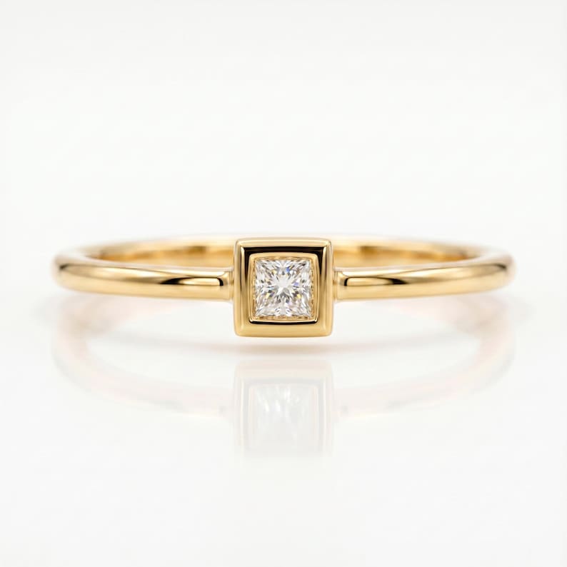 A gold ring with a bezel-set square princess cut diamond centered on a white reflective surface.