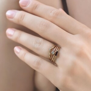 A hand wearing a stack of four delicate gold rings with different gemstones including a V-shaped band, a tiny diamond band, a square diamond band, and a dark stone band.
