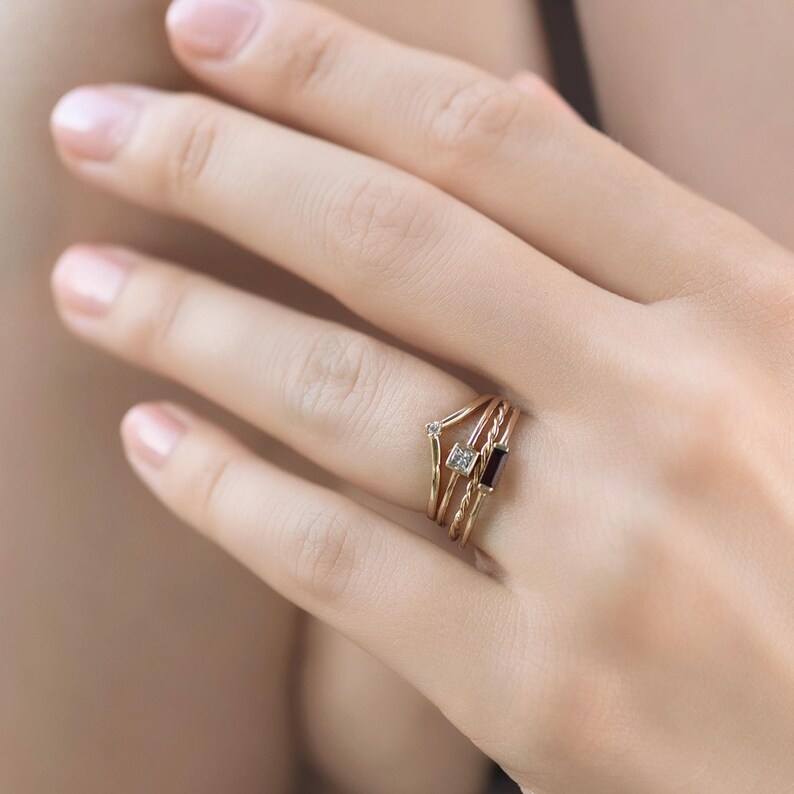 A hand wearing a stack of four delicate gold rings with different gemstones including a V-shaped band, a tiny diamond band, a square diamond band, and a dark stone band.